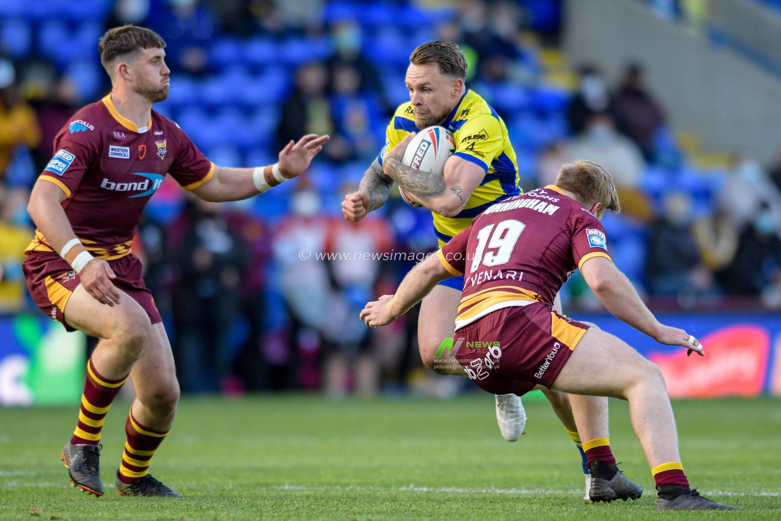 Blake Austin (6) of Warrington Wolves is tackled by James Cunningham (19) of Huddersfield Giants and Joe Greenwood (15) of Huddersfield Giants